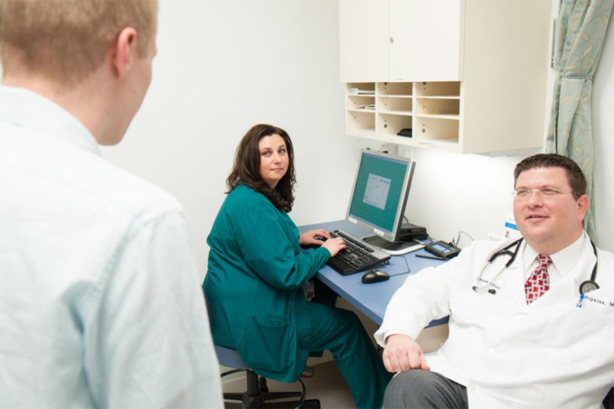 Nurse in green scrubs, sitting at computer and looking back at doctors in the office.