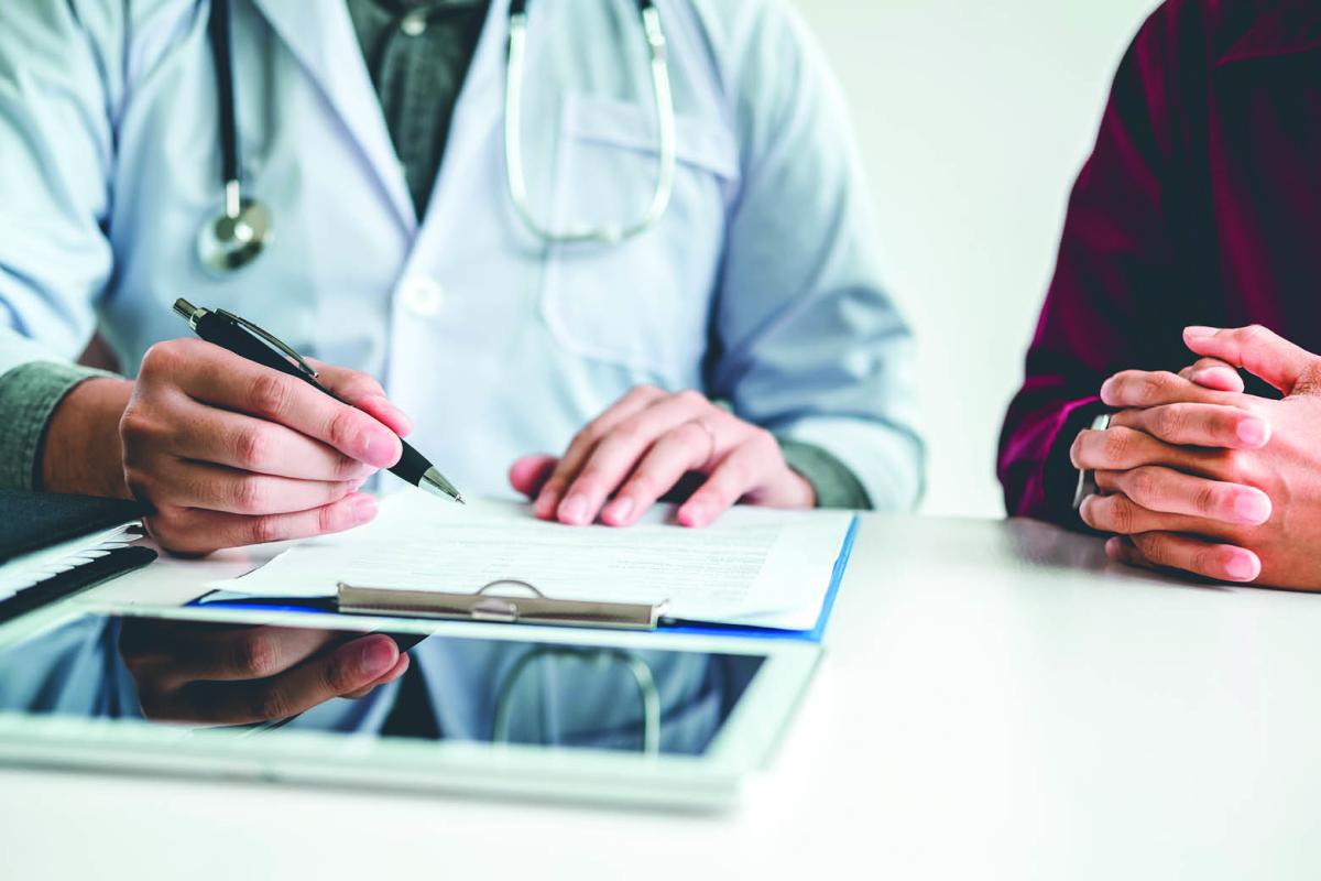 A physician writing on a clipboard on a table by a patient's folded hands