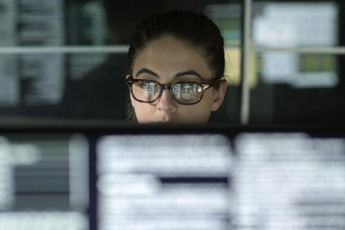 Woman in glasses working at a computer in a darkened room with blurred computer monitors in the foreground.