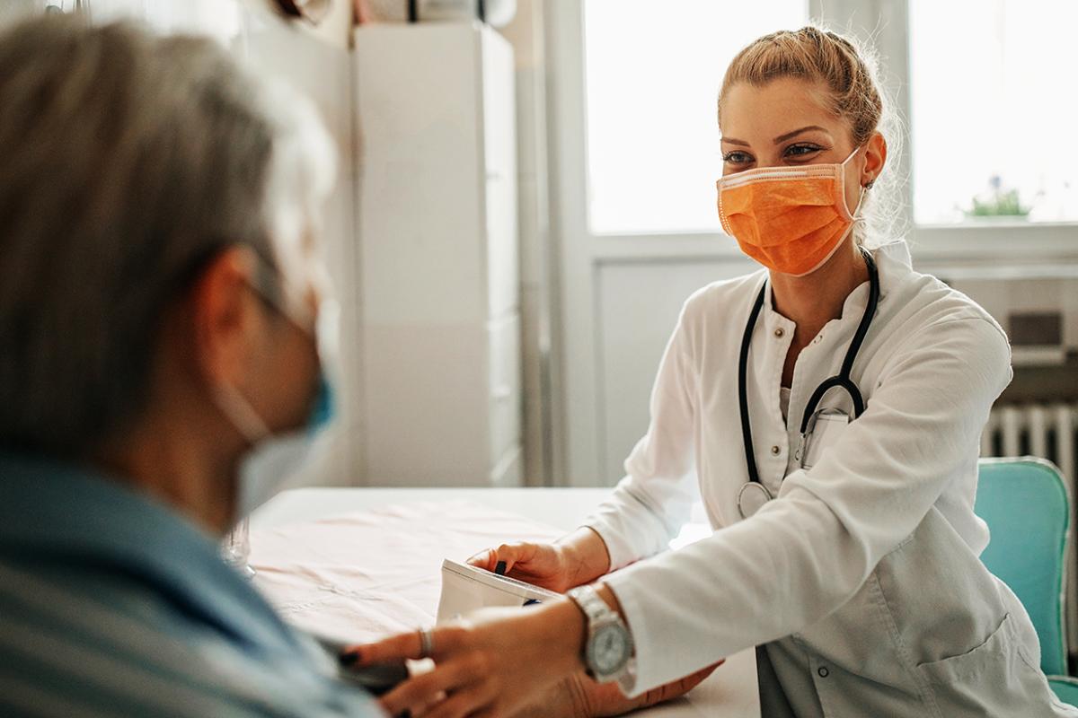 Physician in orange mask sitting down and measuring patient's blood pressure.