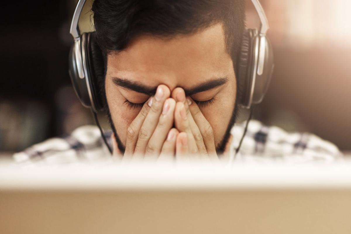 Man in front of computer monitor with eyes closed, hands clasped and wearing headphones. Tight shot.