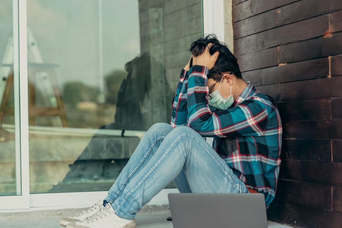 Person sitting outside of a building, wearing a mask, holding their head in their hands.