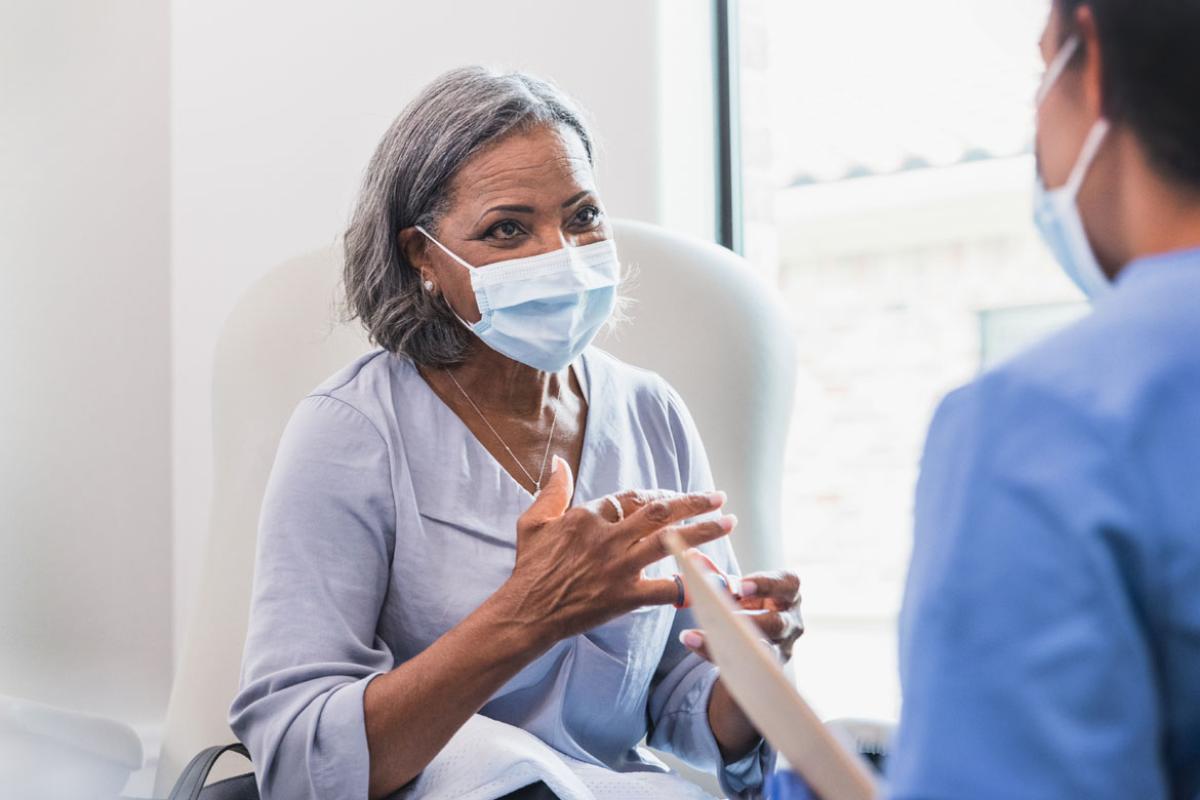 In the background a woman speaking with her doctor in the foreground, both wearing masks.