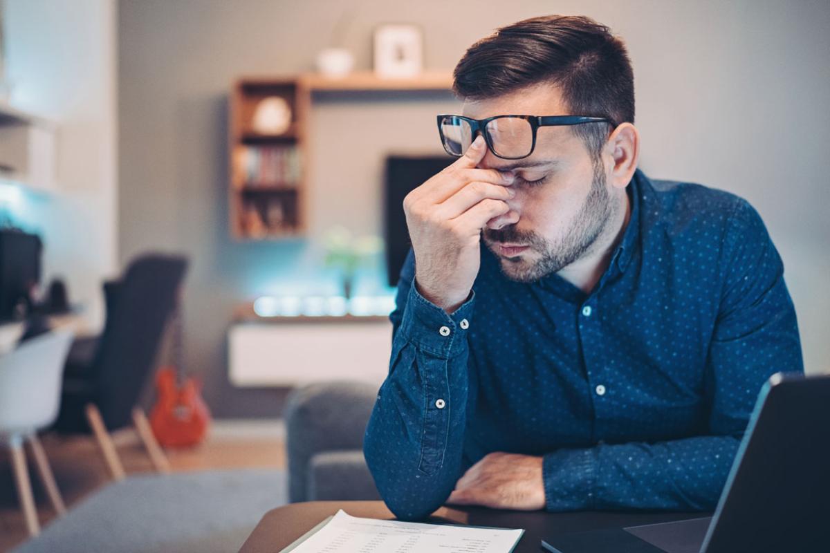 Man sitting at a desk with eyes closed, pinching bridge of his nose with glasses pushed to his forehead.