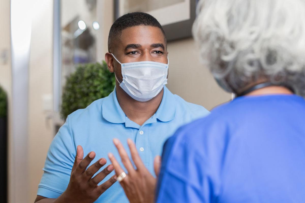 Man in a mask speaking with someone in the foregoround.
