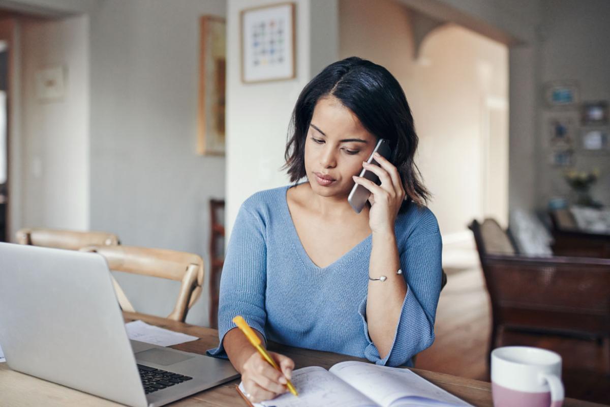 A woman sitting as a desk taking notes while talking on her smartphone in from of her laptop.