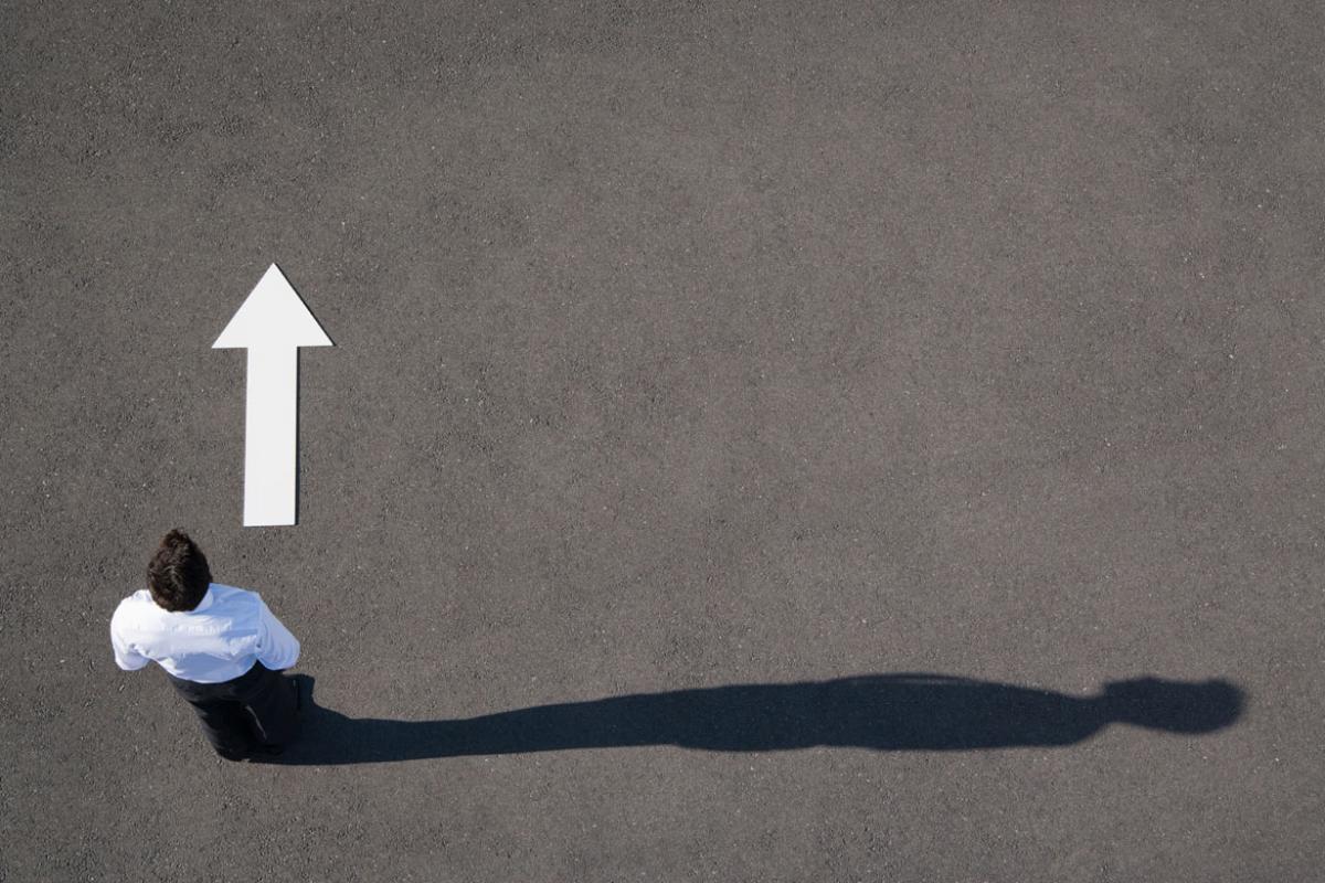 Overhead of a white arrow on the left side being approached by a person casting a long shadow which extends almost entirely right.
