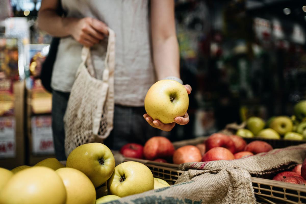 Close-up of a person's torso as they're holding a yellow apple, standing behind bins of yellow and red and yellow apples.
