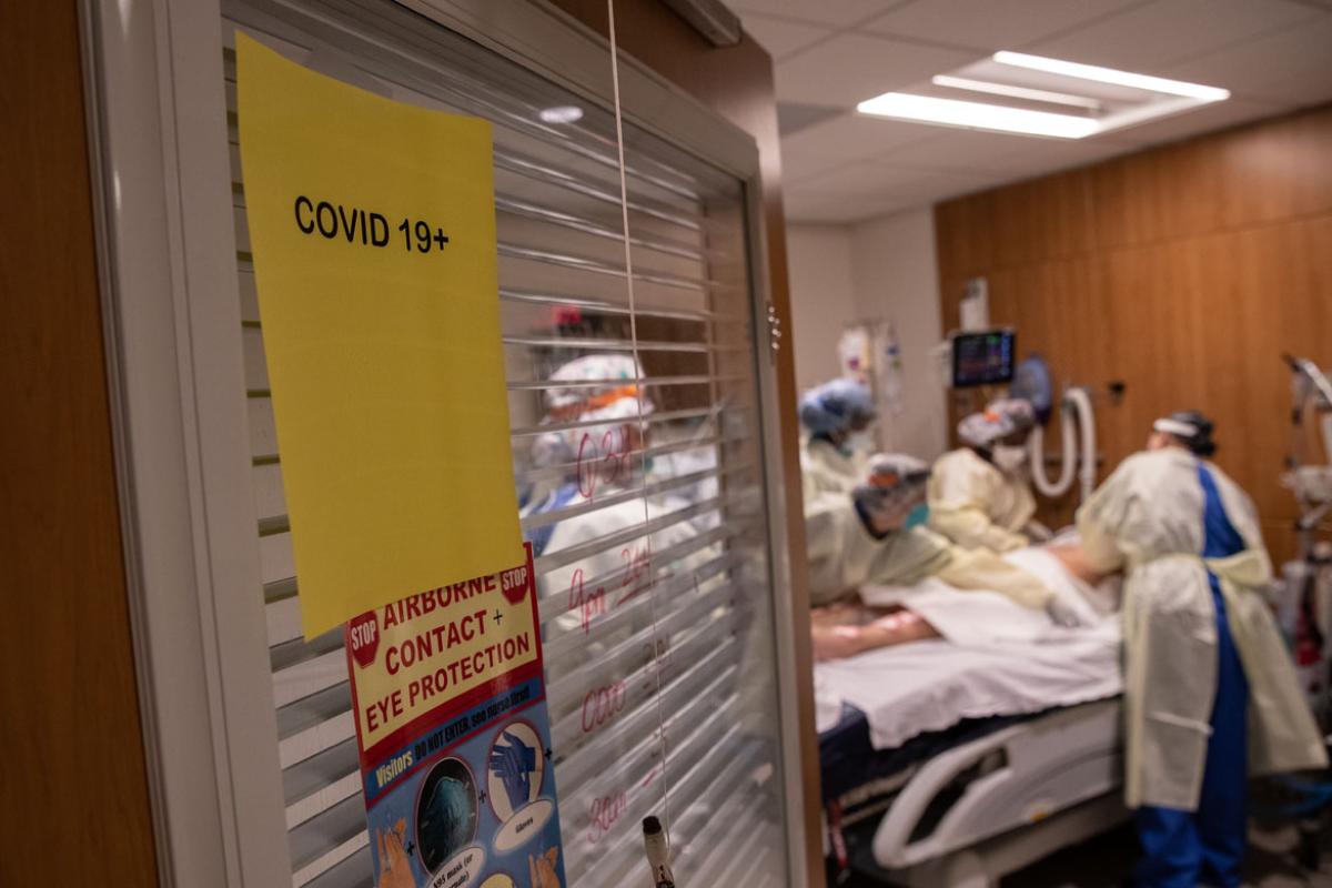 Five doctors and health care workers in full scrubs and PPE working on a patient in a hospital bed in a COVID-19 ward.