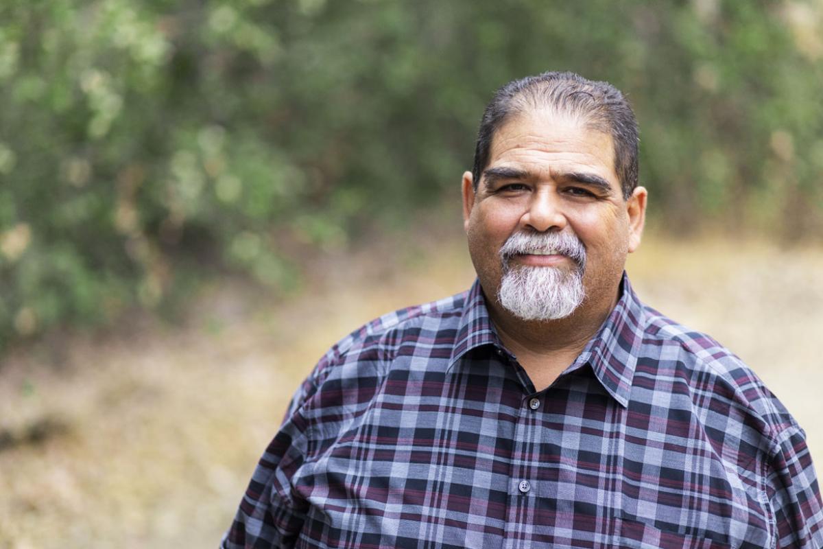 A LatinX man, with a greying goatee beard and short hair, wearing a blue plaid shirt, shown in torso smiling at the camera, outdoors on a hiking path.