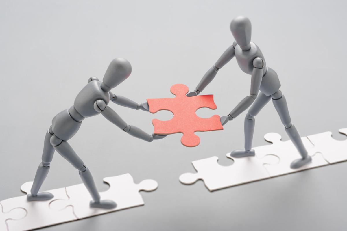 Two grey artist mannequins facing each other, standing on a line of white jigsaw pieces, holding a red jigsaw piece as if to place it to connect the line.