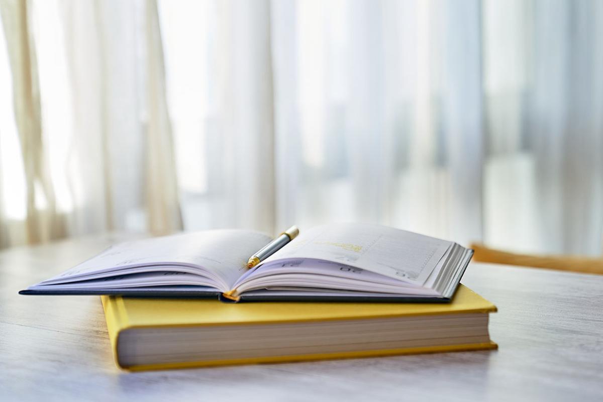 An open journal and pen sitting on top of a yellow covered book, with white curtains in the back ground.