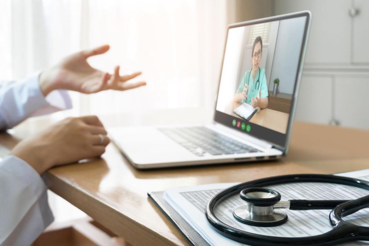 A physician sitting at a desk having a teleconference with another health professional via a laptop, with a stethoscope and clipboard on the right side of the desk.