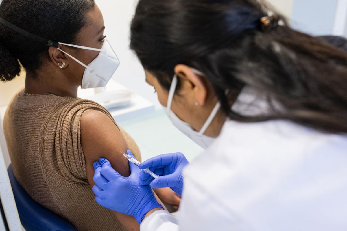 A young African-American woman wearing a mask receiving a shot from a health care worker wearing gloves and a mask.