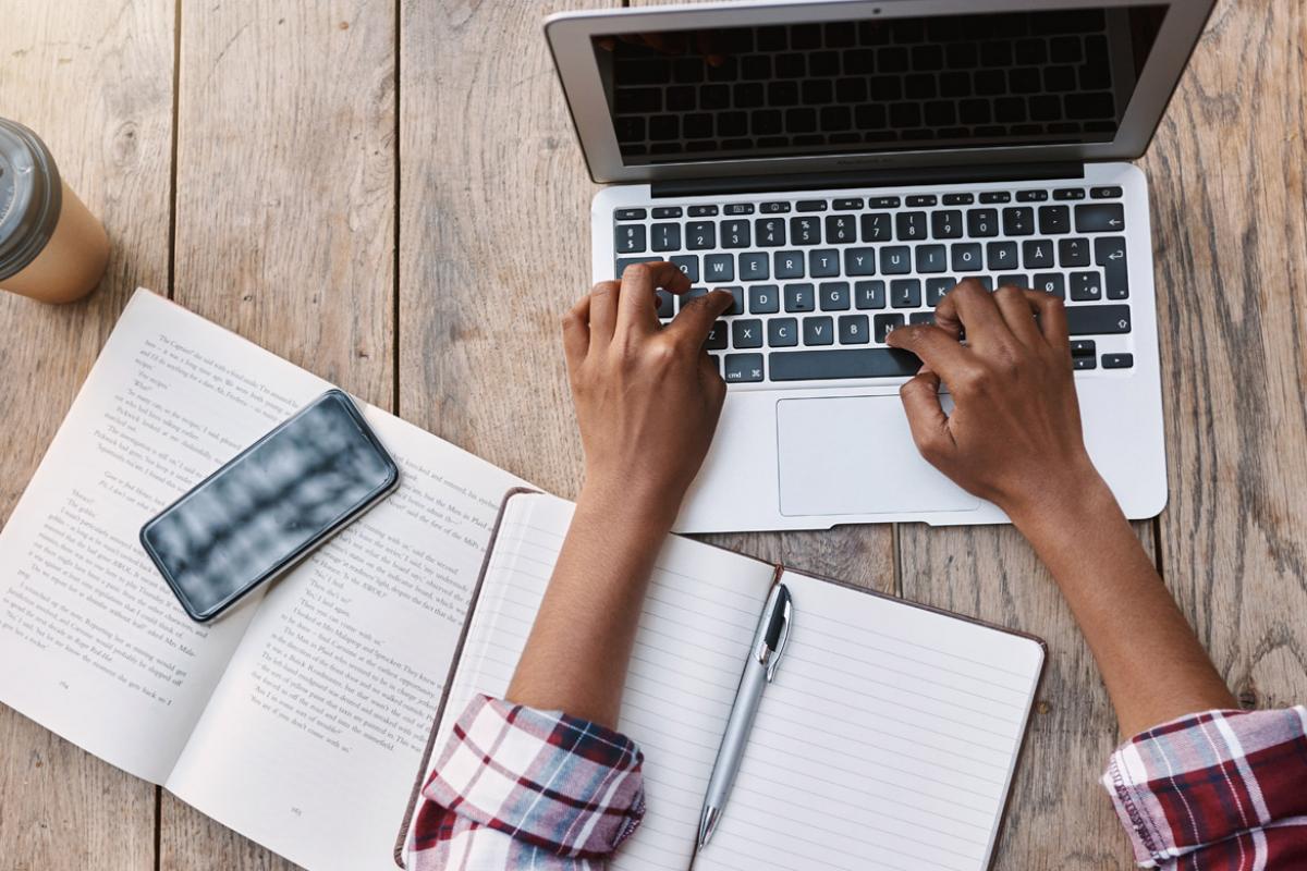 Close up of person's hands typing on laptop keyboard, with a book, notepad, pen, cell phone and beverage cup on the table