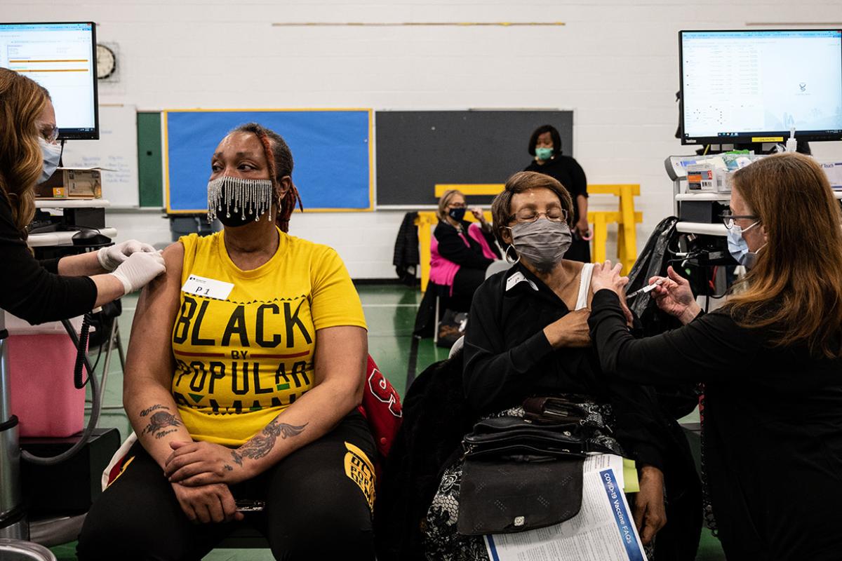 Two women wearing masks and receiving vaccinations at a school or communinity center.