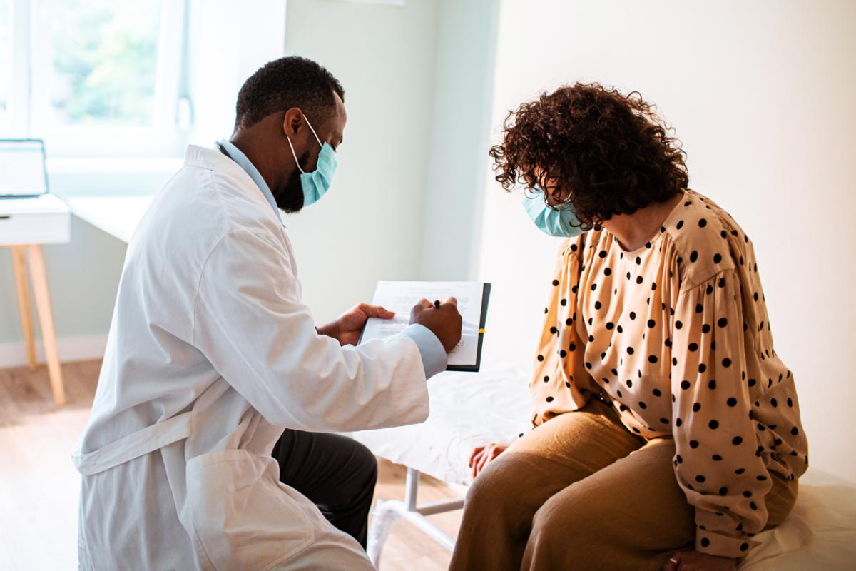 Physician going over examination notes with patient, both wearing masks. 