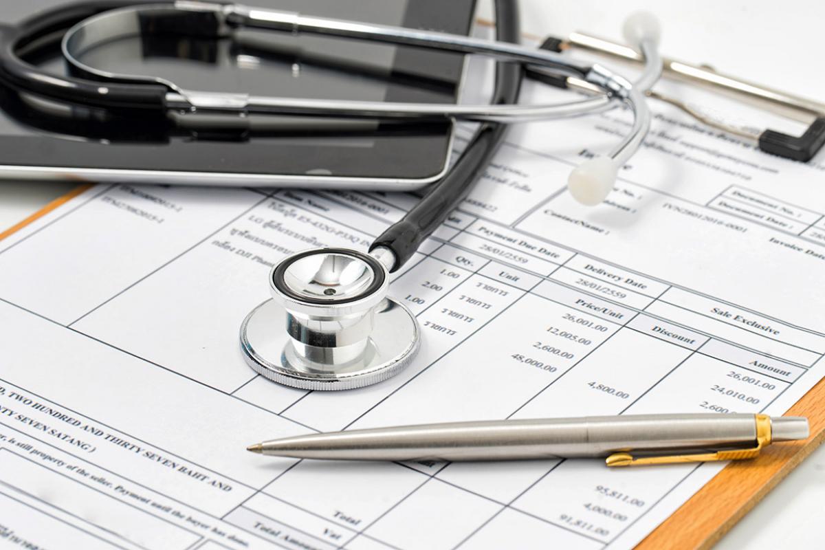 Stethoscope, laptop and pen on top of a medical bill attached to a clipboard