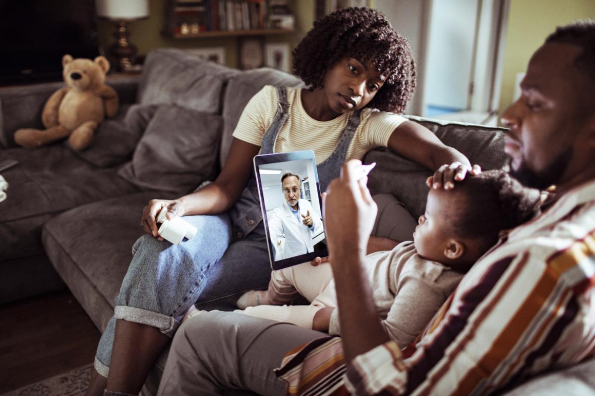 Father and mother sitting on couch with their ill baby having a telehealth call with a physician; the father taking the baby's temperature and the mother feeling the baby's forehead