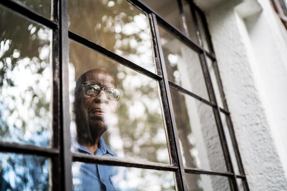 Exterior shot of an older man with a pensive look on his face peering out a block of windows