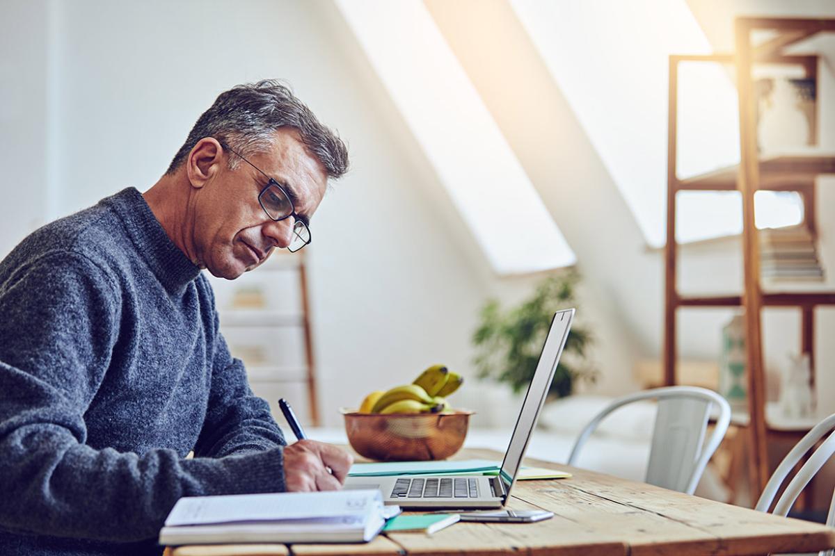 Man sitting at table writing down notes with an open laptop and bananas in a bowl on top of table
