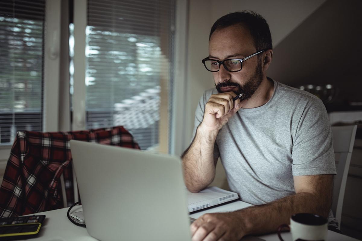 Man at dining room table sitting down with a notebook and pen to watch something on a laptop 