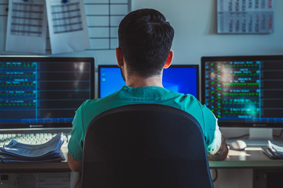 Back view of a health care professional at a desk reviewing patients' records on various computer monitors
