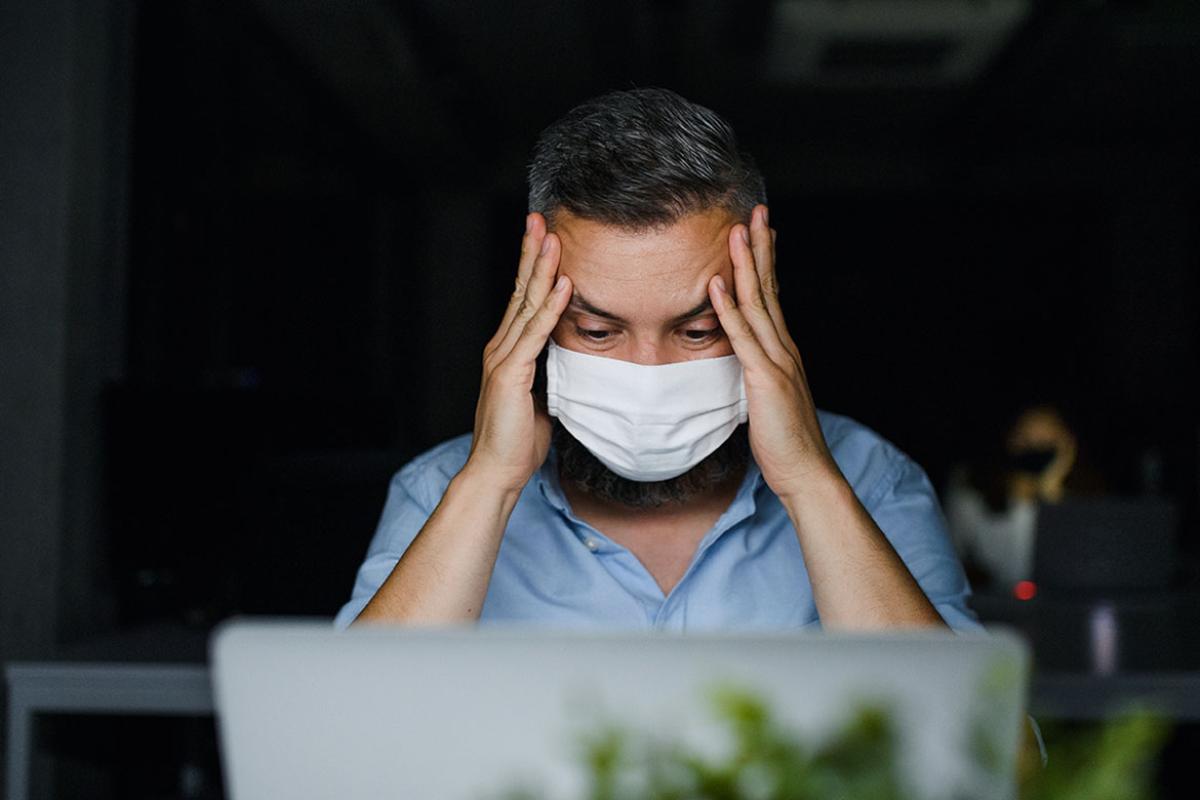 Masked person in front of a laptop with head in hands