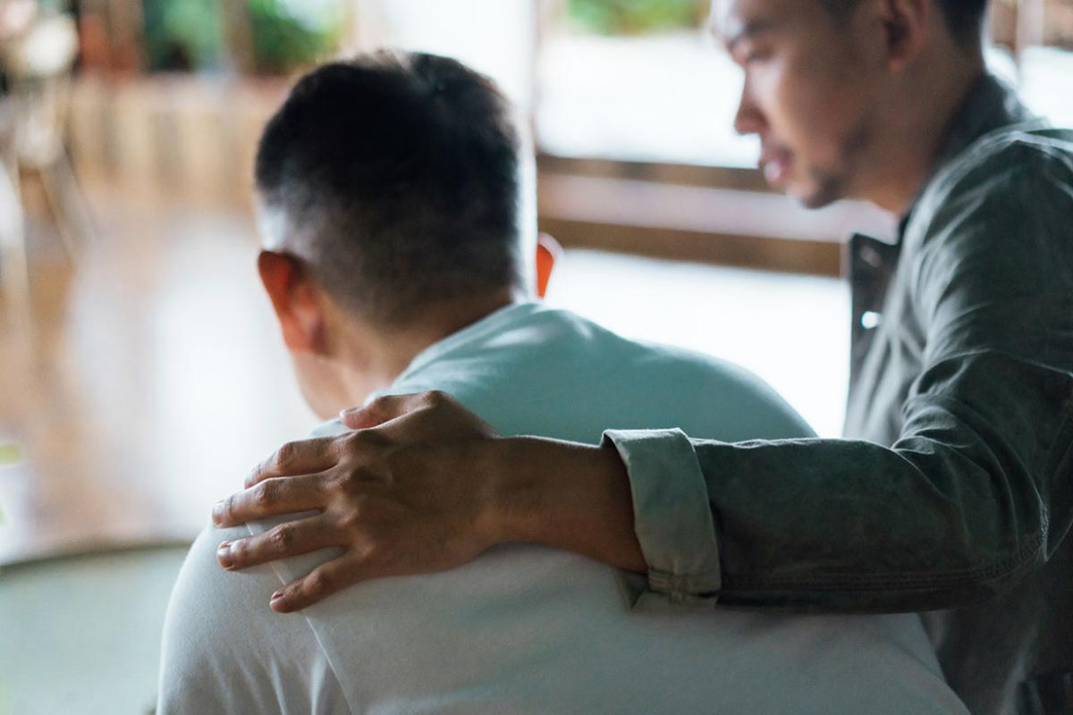 Two people sitting together,  one putting hand on another's shoulder