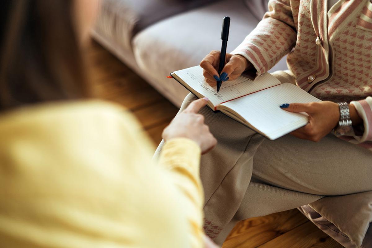 Two people in armchairs are sitting and one is writing in a notebook