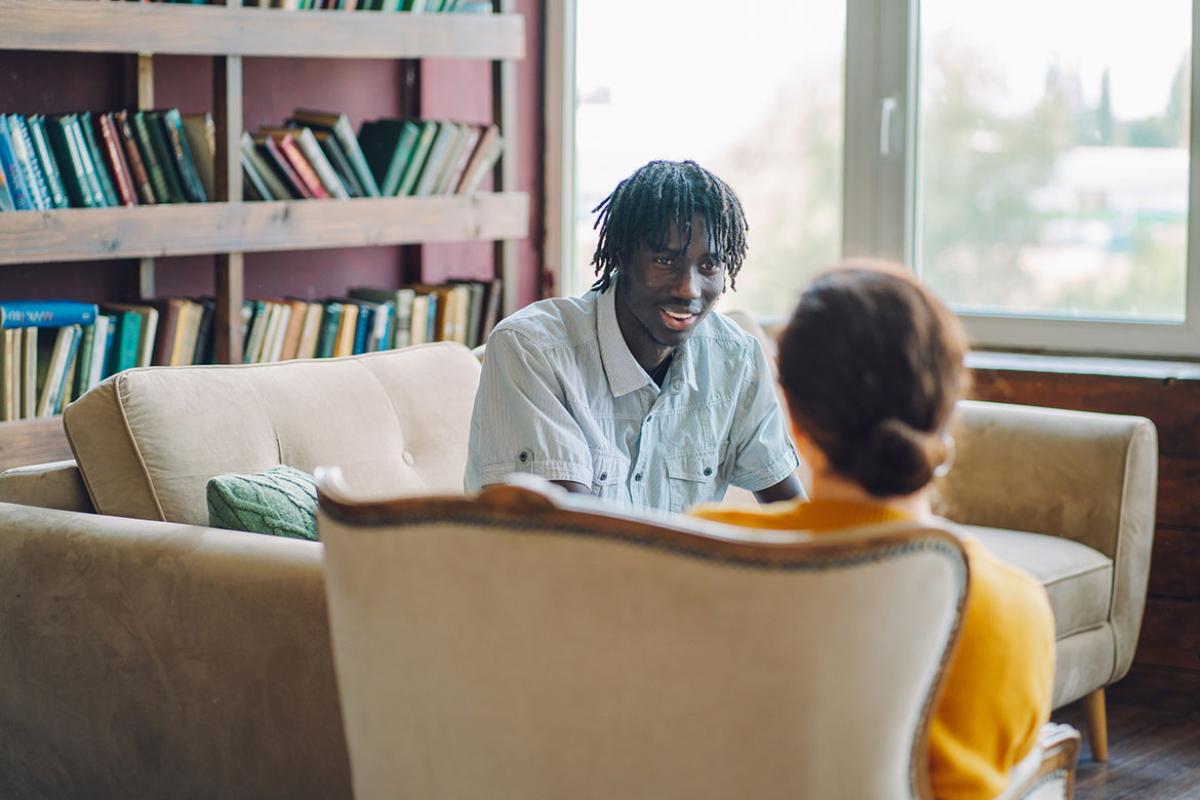 A young man sit on a sofa in an office speaking to another individual