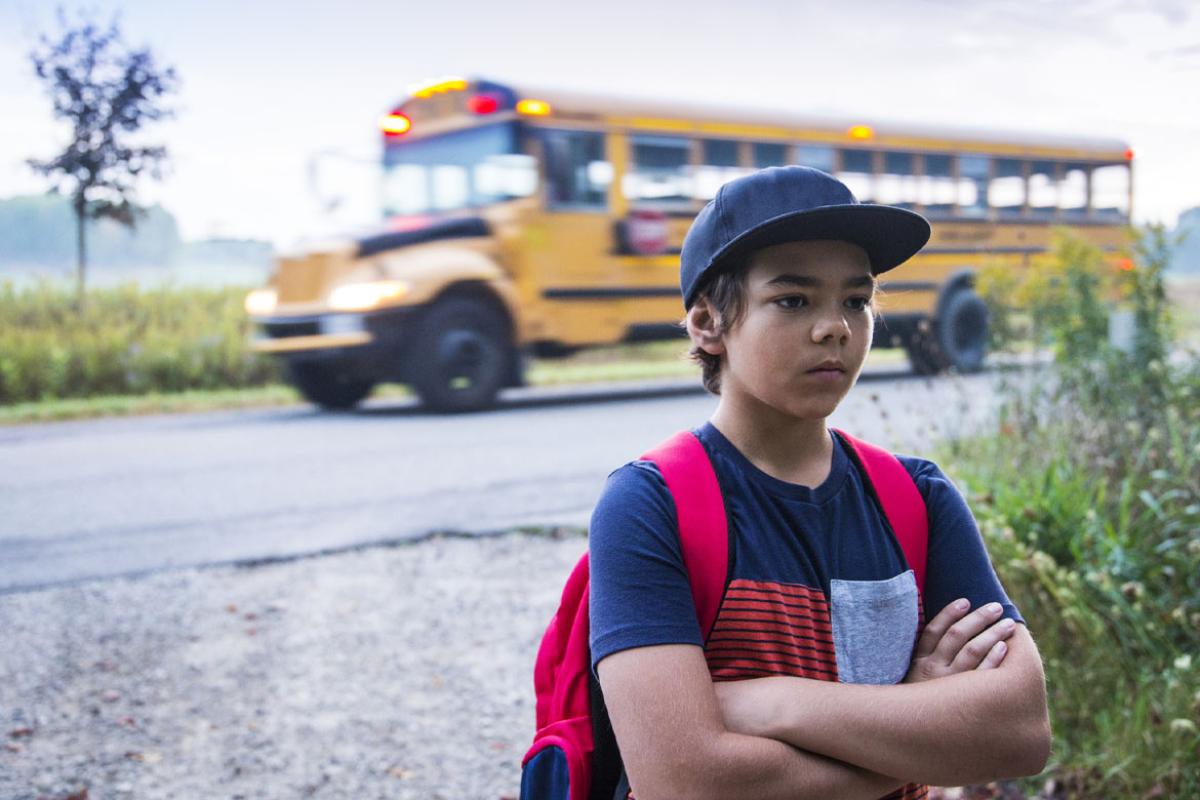 Child wearing a backpack with arms crossed, bus in the background
