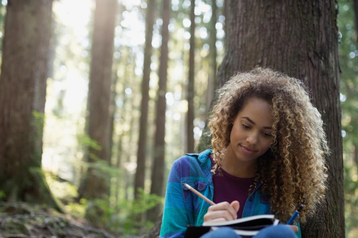 A woman outside in a forest writing in a journal in pencil sitting against a tree.