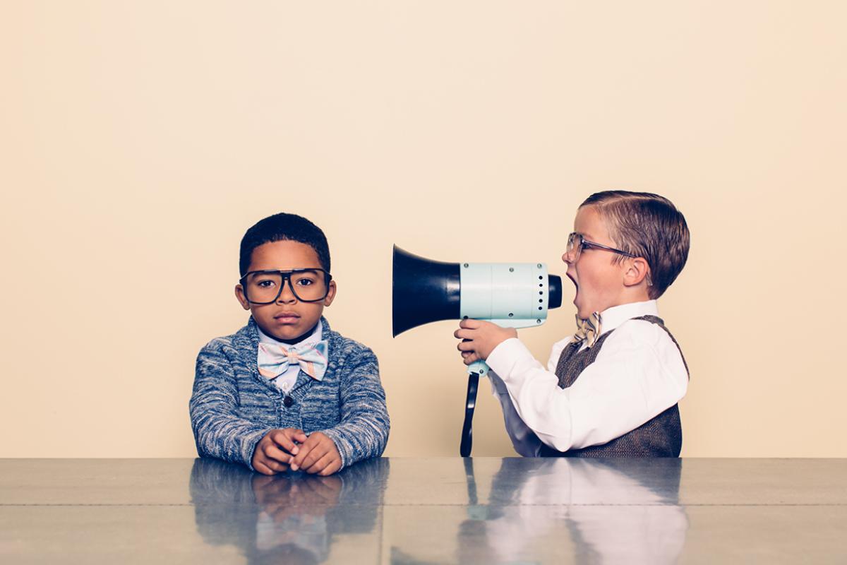 2 boys wearing bowties sitting at a table. One is shouting through a loudspeaker at the other.