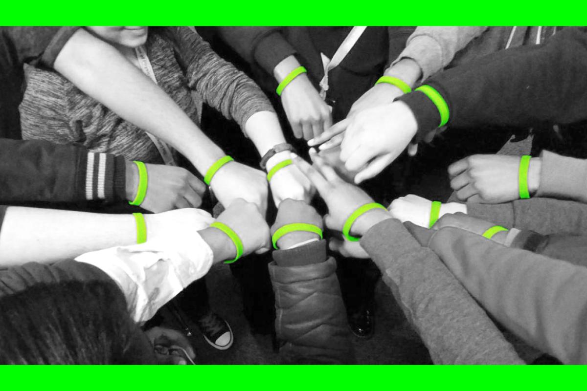 A group of teens putting their hands in a circle all wearing a neon green wristband.